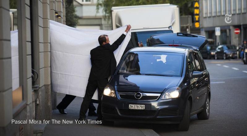 A white blanket is held up by a man in a suit in order to cover the entrance of an indictee into a car. 